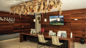 A reception area with a wooden wall, a TV screen, and a desk with chairs. - Luxury Condo in PGA Golf Resort | Bahia Principe (Akumal)