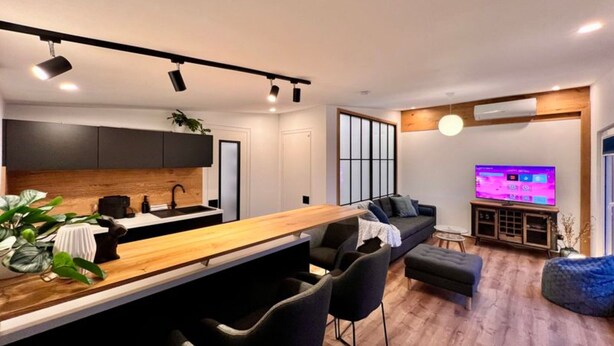 Una cocina moderna con encimera de madera y sillas negras, un área de comedor con sofá y reposapiés, y un televisor montado en la pared.