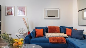 Un salón con un sofá azul, cojines naranjas y una mesa de centro de cristal. Hay dos cuadros en la pared y un jarrón con plantas secas sobre una mesita.