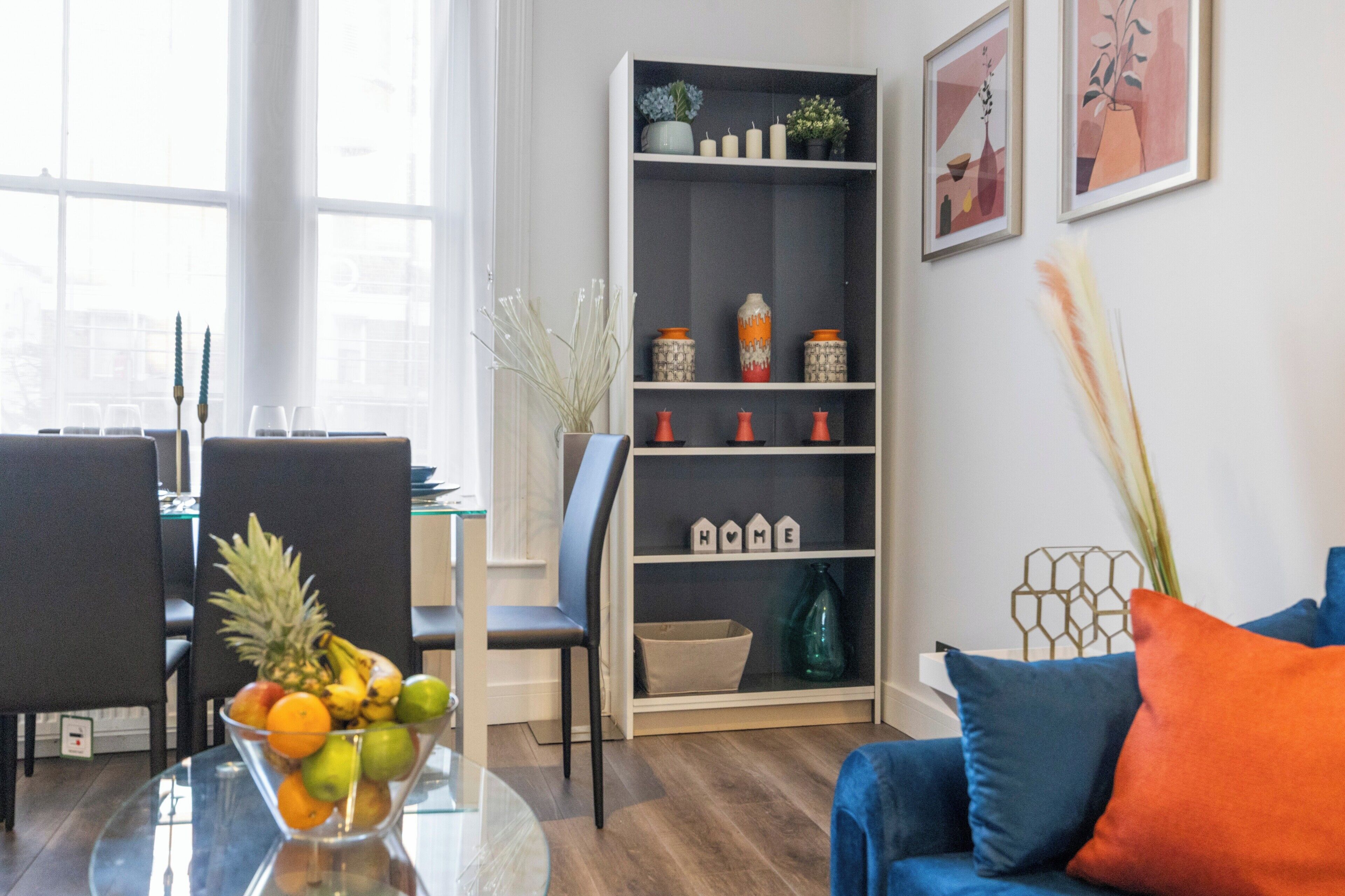 A modern living room with a glass coffee table, a blue sofa, and a bookshelf with decorative items.