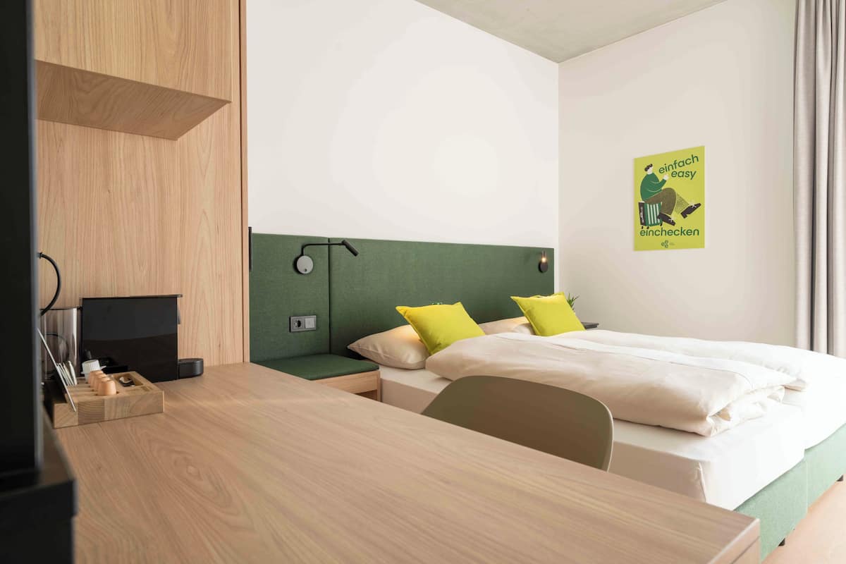A modern hotel room with a wooden desk, a bed with green headboard, and a poster on the wall.