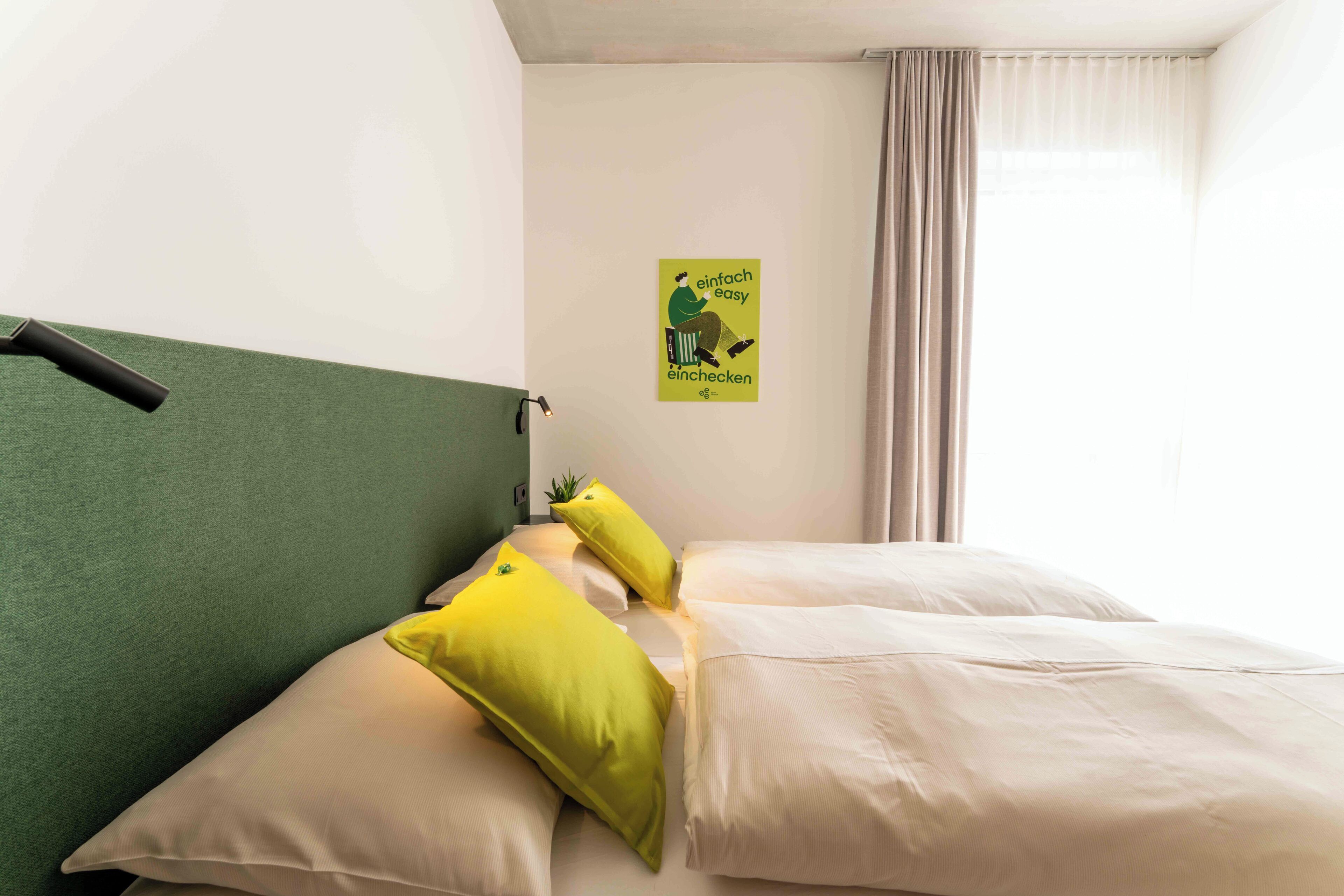 Une chambre d’hôtel avec une tête de lit verte, deux oreillers jaunes, une couvre-lit beige et une affiche accrochée au mur.