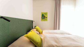 Une chambre d’hôtel avec une tête de lit verte, deux oreillers jaunes, une couvre-lit beige et une affiche accrochée au mur.