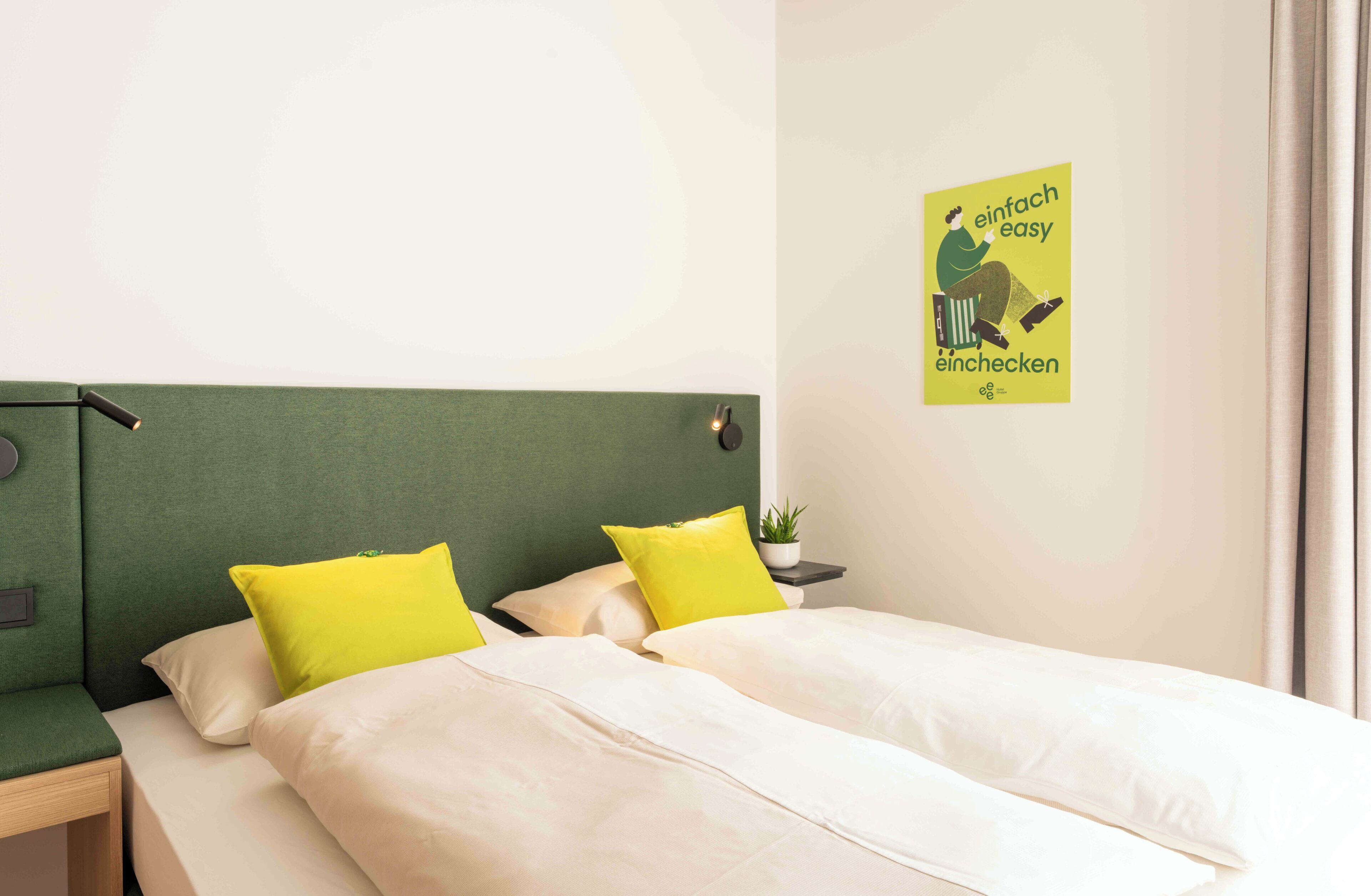 A hotel room with a green headboard, two beds with white and yellow bedding, a small bedside table with a lamp, and a poster on the wall.