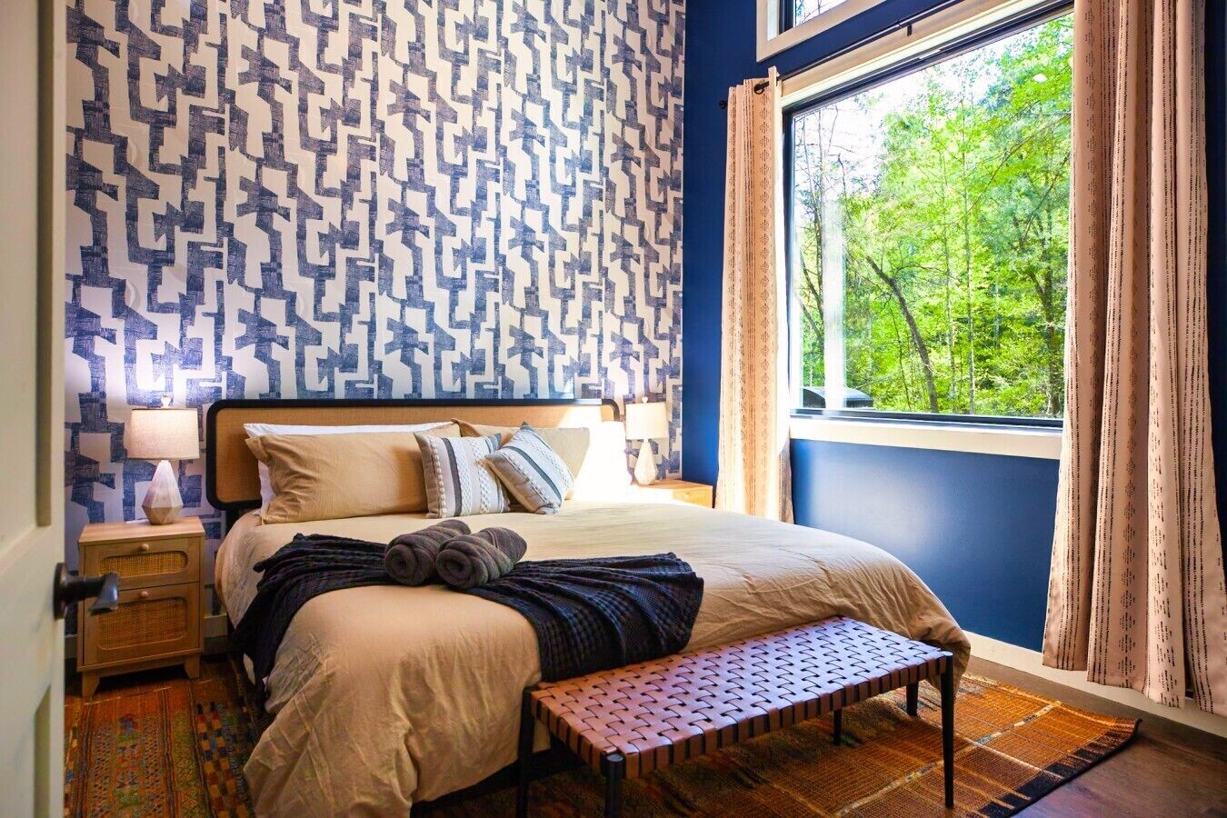 A bedroom with a patterned wall, a bed with pillows, a bench, a nightstand, a lamp, and a view of trees through a window.