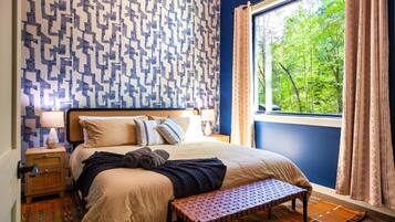 A bedroom with a patterned wall, a bed with pillows, a bench, a nightstand, a lamp, and a view of trees through a window.