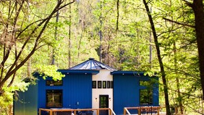 Une cabane bleue avec une terrasse en bois et un toit en métal, entourée d’arbres.