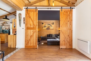 A rustic interior with exposed wooden beams, a sliding barn door entrance, a dark wall with a fan, and a sofa with patterned cushions. - Tropical Escape in Dordogne (Le Fleix)