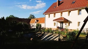 Une maison à deux étages, avec un toit recouvert de tuiles rouges, un porche à l’avant et un petit jardin avec des marches menant à l’entrée.
