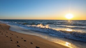 Ranta, jossa näkyy jalanjälkiä, jotka johtavat veteen, aallot lyövät rantaan ja aurinko laskee taivaanrannassa.