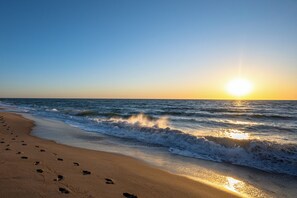 Ein Strand mit Fußabdrücken, die zum Wasser führen, Wellen, die an das Ufer brechen, und einer untergehenden Sonne im Hintergrund.