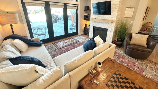 Een moderne woonkamer met een bank, salontafel en open haard. Een televisie is aan de muur bevestigd en er is een groot raam met zonwering.