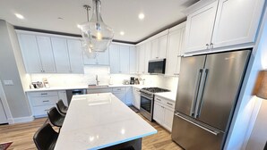 Een moderne keuken met witte kastjes, een roestvrijstalen koelkast, een groot eiland met stoelen en een hanglamp.