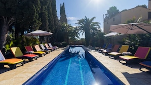 A swimming pool with a tiled edge, surrounded by lounge chairs and umbrellas, with trees and a building in the background. - Masia Notari Casabona (Vilanova I la Geltru)