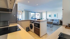 Een moderne keuken met een ingebouwde wasmachine en oven, een zithoek met een bank, en stadszicht door grote ramen.