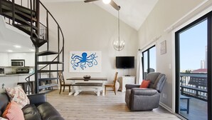 A modern living room with a black leather sofa, a gray armchair, a dining area with a white table, a black TV, and a large window with a city view. - NEW LISTING!! Regatta Condos 304C - Beachfront Resort w/Pool (Gulf Shores)