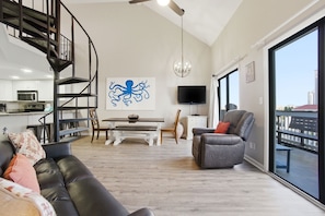 A modern living room with a black leather sofa, a gray armchair, a dining area with a white table, a black TV, and a large window with a city view. - NEW LISTING!! Regatta Condos 304C - Beachfront Resort w/Pool (Gulf Shores)