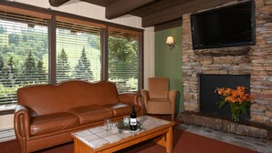 A living room with a leather sofa, a wooden coffee table, a stone fireplace, a television mounted on the wall, and a window with blinds.