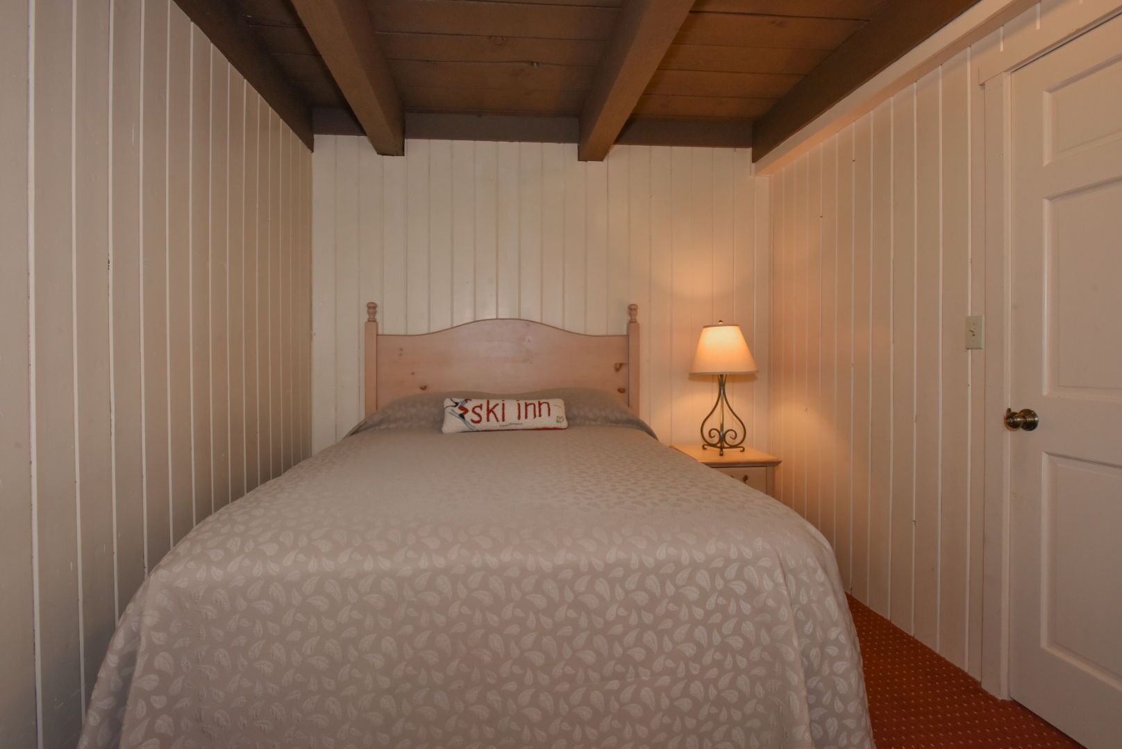 A bedroom with a wooden headboard, a bed with a floral-patterned bedspread, a nightstand with a lamp, and a door with a brass doorknob.