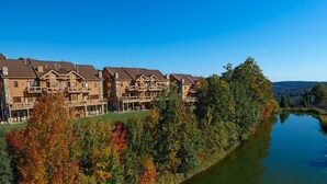 Une rangée d’immeubles imposants, à plusieurs étages et dotés de balcons, se dresse au bord d’un lac paisible, entourée d’arbres aux couleurs automnales.