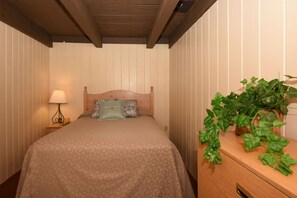 Une chambre à coucher avec une tête de lit en bois, un lit recouvert d’une literie à motifs, une lampe de chevet et une plante en pot posée sur une commode en bois.