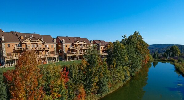Une rangée d’immeubles imposants, à plusieurs étages et dotés de balcons, se dresse au bord d’un lac paisible, entourée d’arbres aux couleurs automnales.
