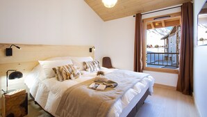 A bedroom with a wooden ceiling, a large bed with white bedding, a nightstand with a lamp, and a window with a view of buildings and mountains. - Ski aux pieds ! Moderne T4 avec vue imprenable (Alpe d'Huez)