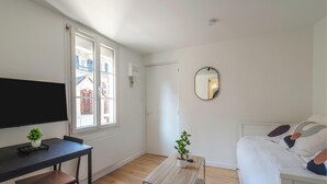 A room with a bed, a TV, a small table with a plant, a mirror, and a window with a view of buildings. - L'Ambroise - Wifi - Centre-ville - Gare (Laval)