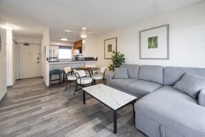 Ein modernes Wohnzimmer mit einem grauen Ecksofa, einem weißen Marmortisch und einem Essbereich mit weißen Stühlen und einem kleinen Tisch.