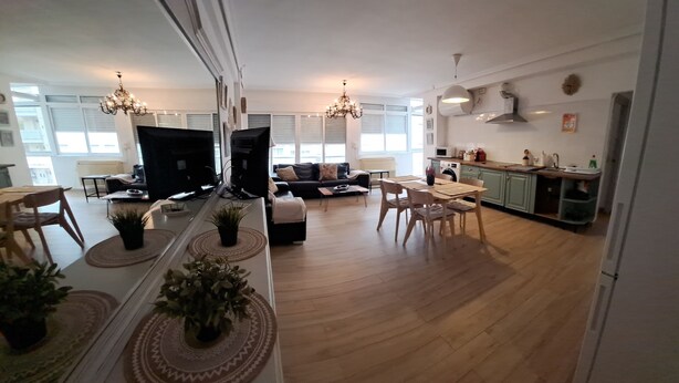 En modern lägenhet med vardagsrum, matplats och kök. Det finns två stora fönster, en kristallkrona och en tv med platt skärm.