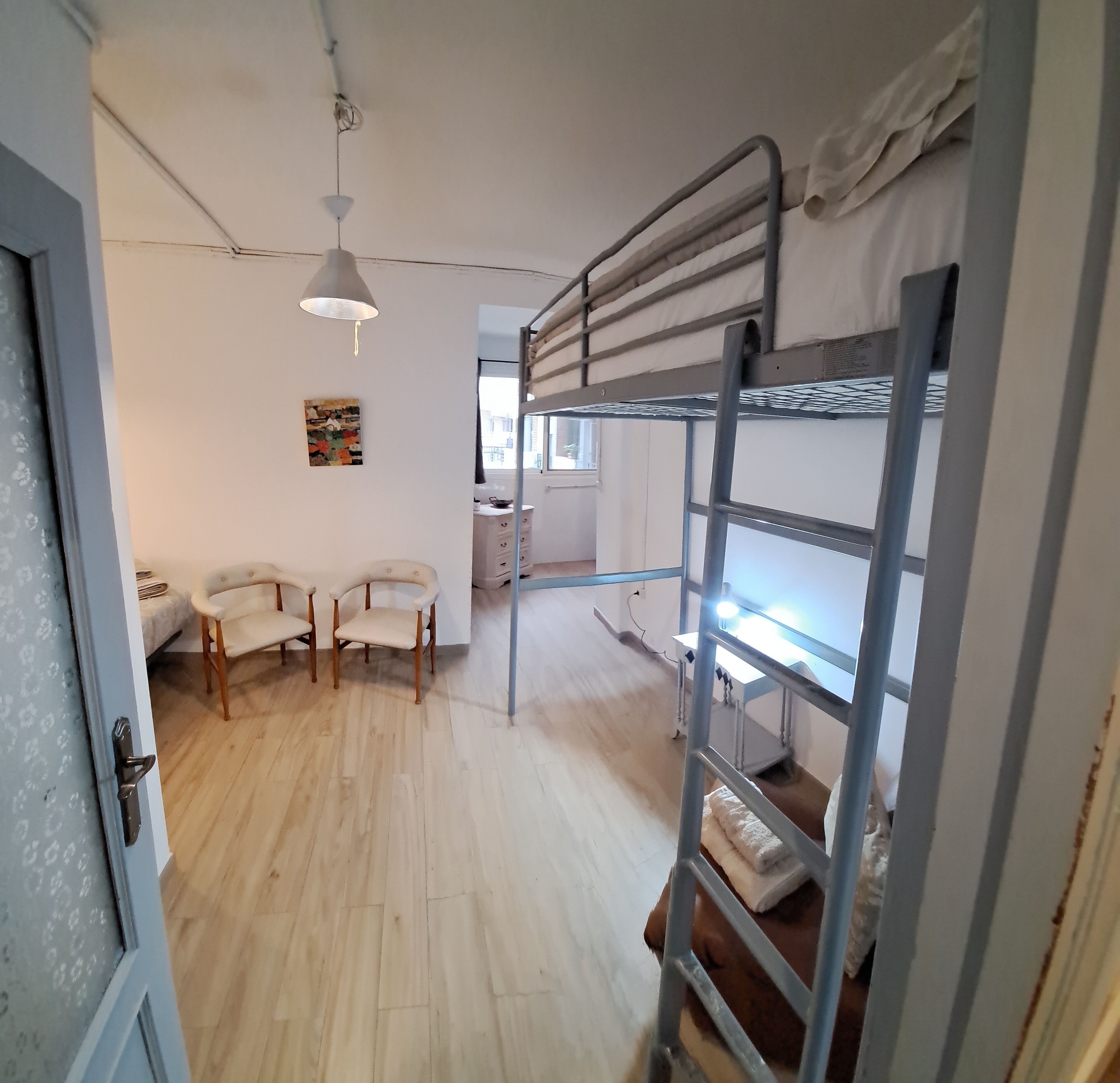 A room with bunk beds, a desk, two chairs, a hanging light fixture, and a window with blinds.