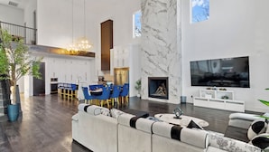 Un salon moderne avec un grand canapé d’angle, un coin repas avec des chaises bleues, une cuisine avec des meubles blancs et une cheminée.