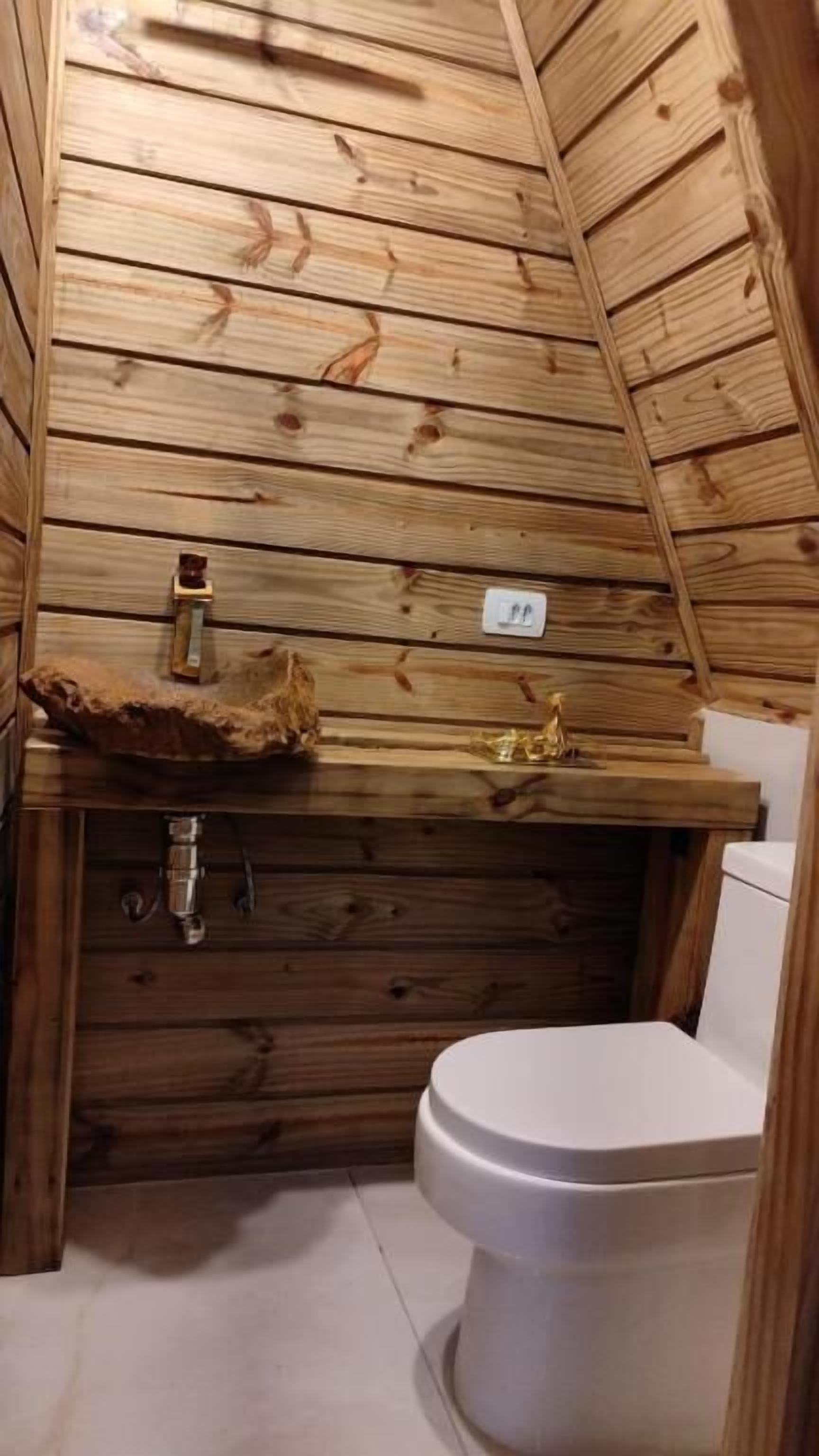 A small, wooden cabin bathroom with a wooden slat ceiling, a sink with a rustic basin, a toilet, and a small shelf with a decorative item.