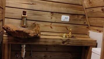 A small, wooden cabin bathroom with a wooden slat ceiling, a sink with a rustic basin, a toilet, and a small shelf with a decorative item.