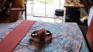 Une chambre mansardée chaleureuse, avec un plafond en pente, un lit recouvert d’une couvre-lit à motifs floraux, une petite table avec un panier, et une vue sur un jardin par une porte vitrée.