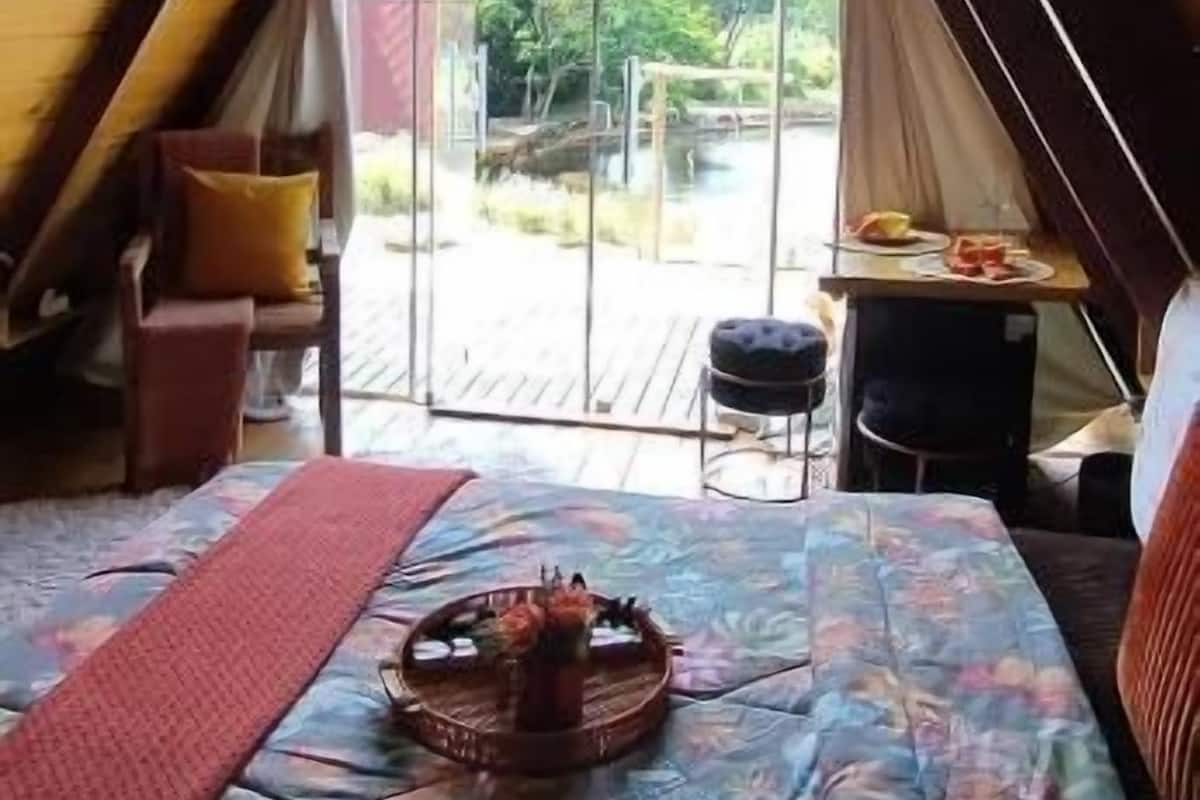A cozy attic bedroom with a sloped ceiling, a bed with a floral bedspread, a small table with a basket, and a view of a garden through a glass door.