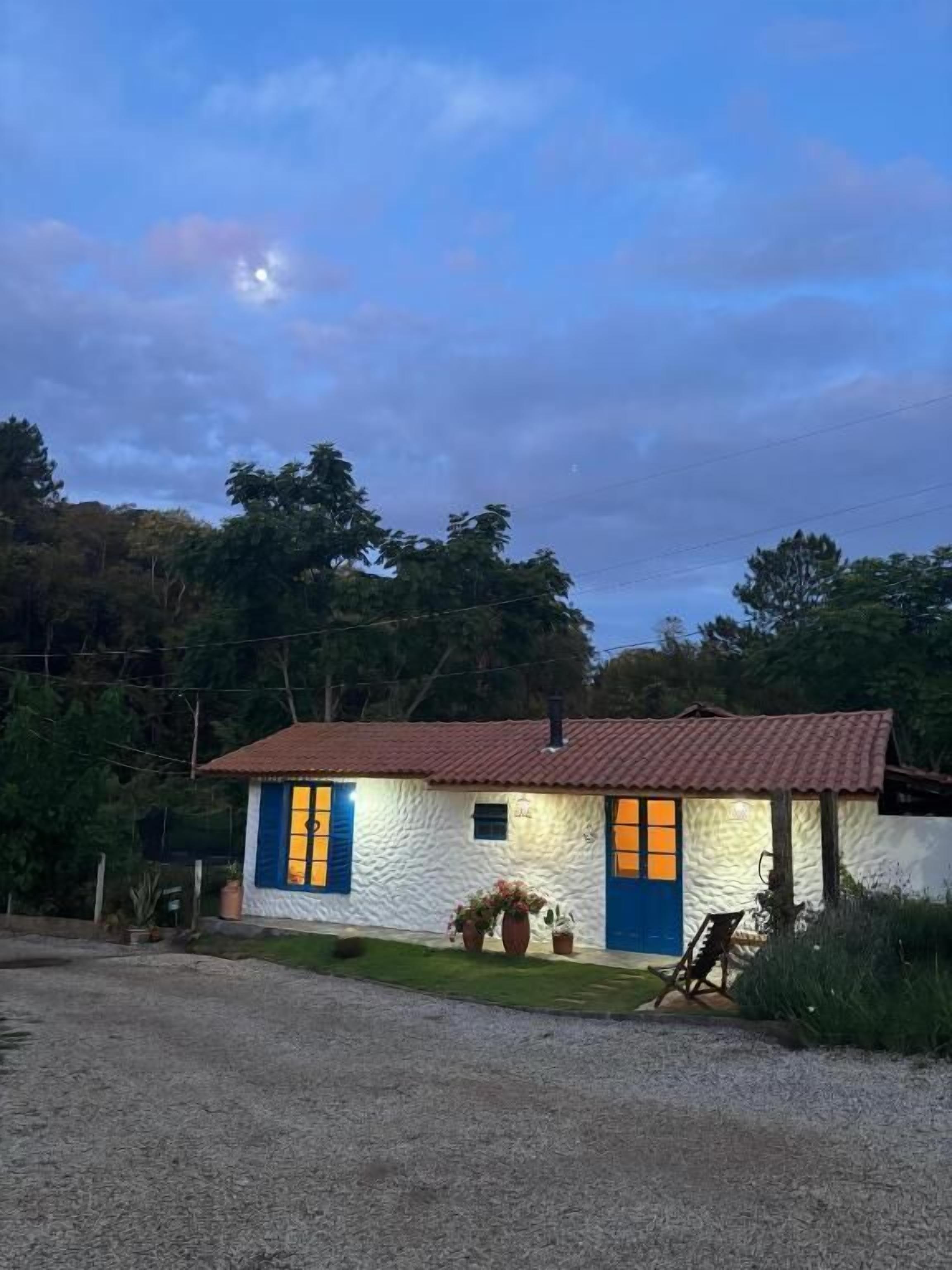 Una casita con techo de tejas rojas, paredes blancas y puertas y ventanas azules. Hay macetas con plantas frente a la casa.