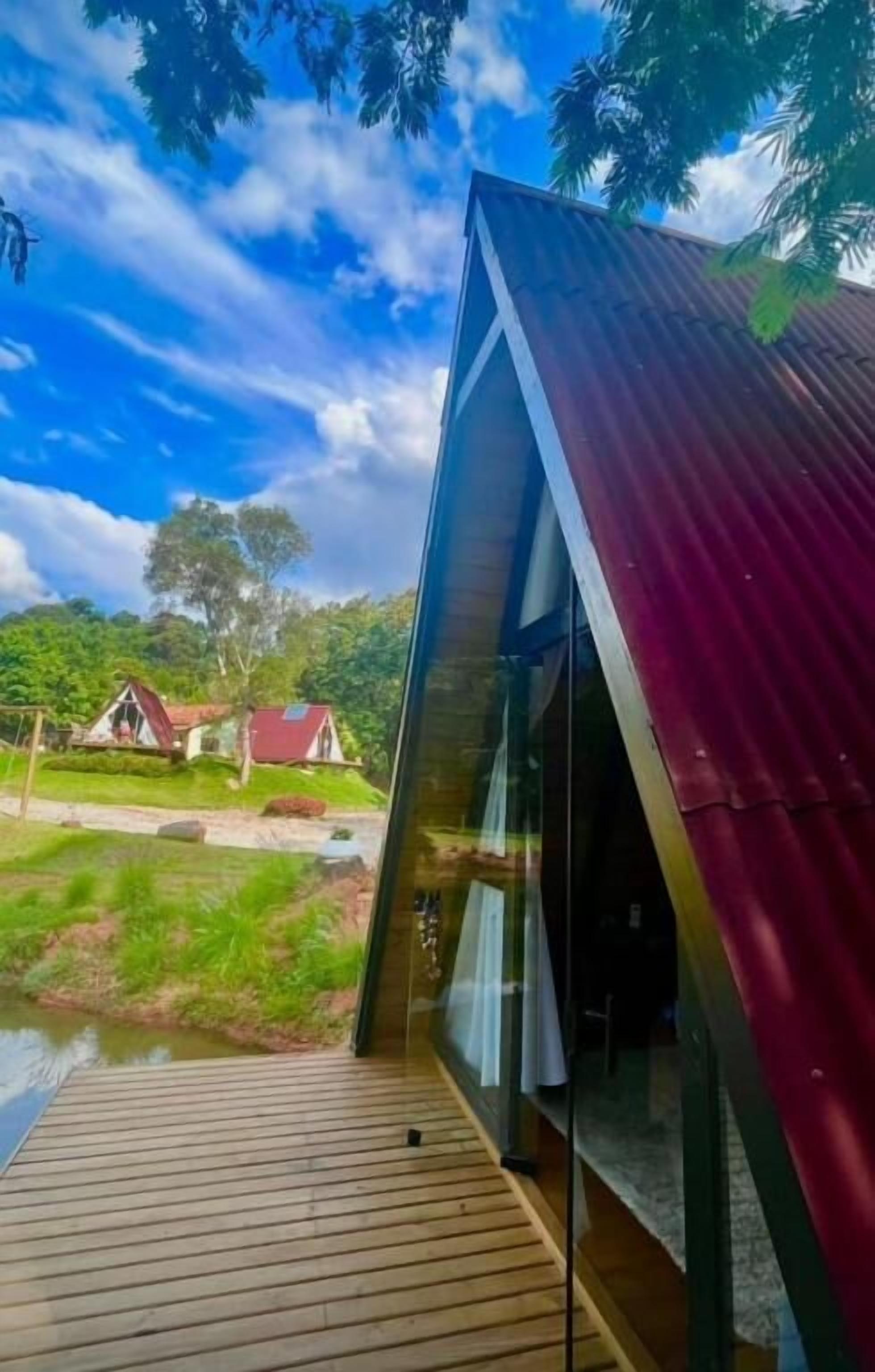 Una estructura moderna de vidrio con techo rojo, terraza de madera y vistas a un paisaje verde exuberante con un cuerpo de agua y edificios a lo lejos.