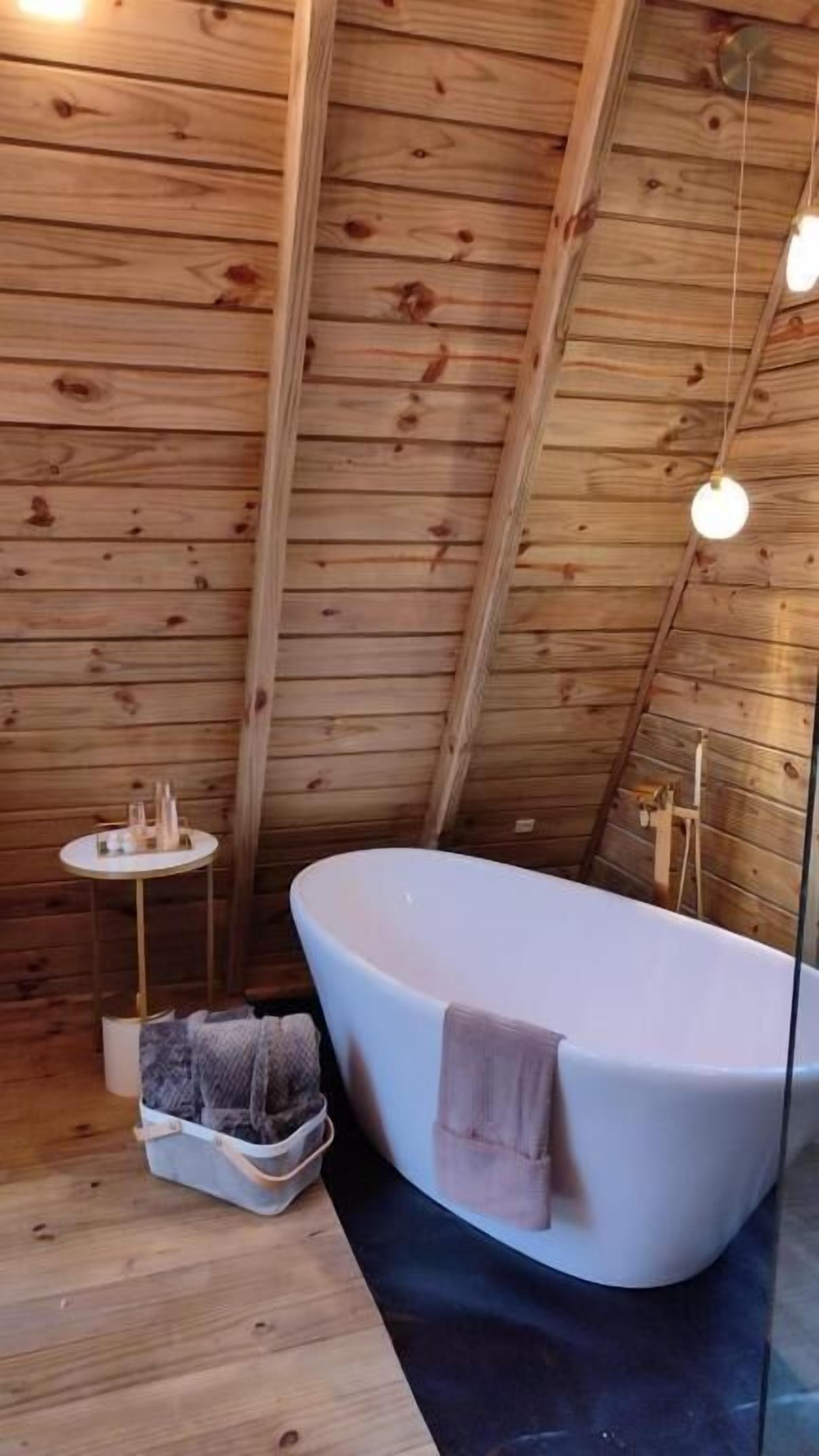 Ein Badezimmer einer Holzhütte mit einer freistehenden Badewanne, einem kleinen Tisch mit Toilettenartikeln und einer Pendelleuchte.