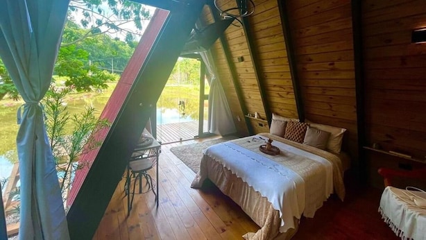 Une chambre à coucher avec un lit, une petite table et une chaise. Il y a une fenêtre avec des rideaux et une vue sur la verdure à l’extérieur.