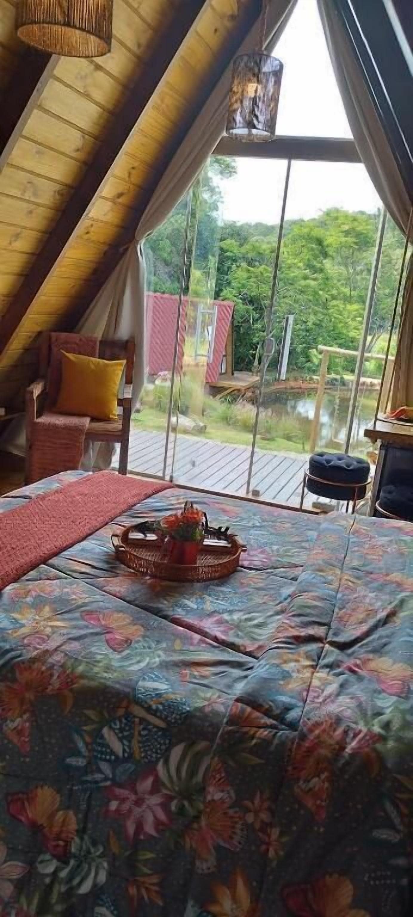 A cozy room with a wooden ceiling, a bed with a floral bedspread, a wicker basket with flowers, and a view of a red building and greenery outside.