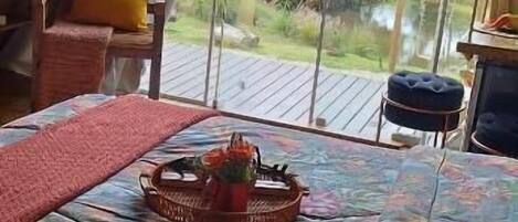Una habitación acogedora con techo de madera, cama con colcha floral, una cesta de mimbre con flores y vistas a un edificio rojo y vegetación.