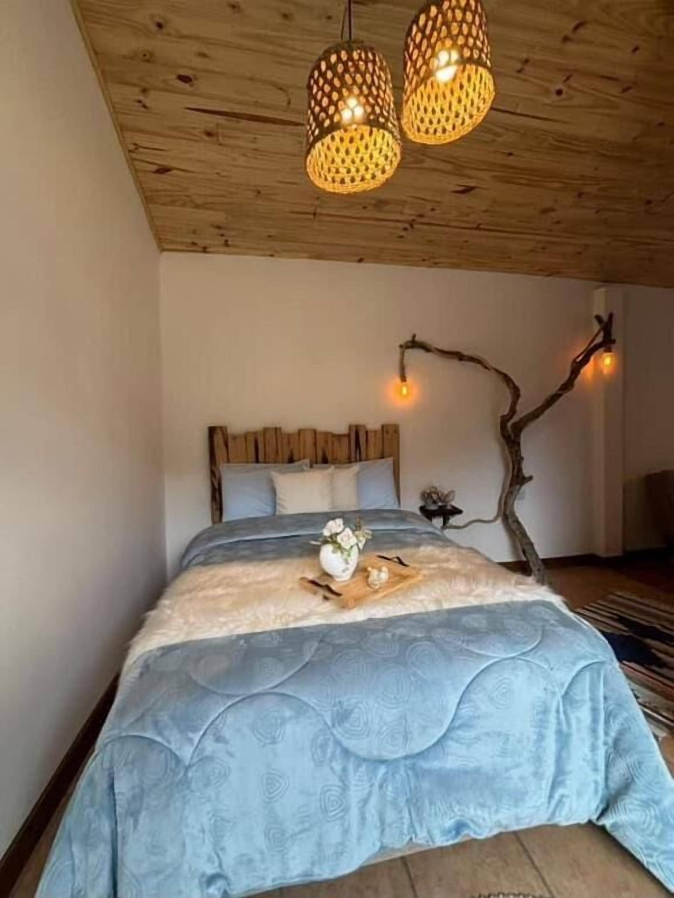 Un dormitorio con techo de madera, una cama con colcha de rayas azules, una decoración de pared con rama de madera y dos lámparas colgantes.