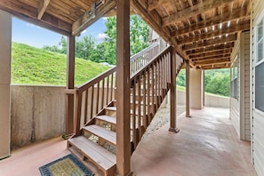A covered outdoor staircase with wooden railings leading to a higher level, surrounded by a concrete wall and a view of a green hillside. - New! Scenic 2BR | Near SDC & Table Rock Lake (Branson)