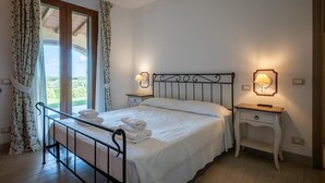 Une chambre à coucher avec un lit, deux tables de chevet, une fenêtre avec des rideaux et une vue sur la verdure à l’extérieur.