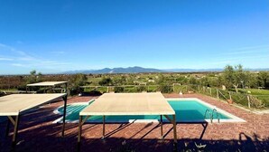 Un espace piscine offrant une vue sur les montagnes lointaines, entouré d’une terrasse en briques et de tables en bois.