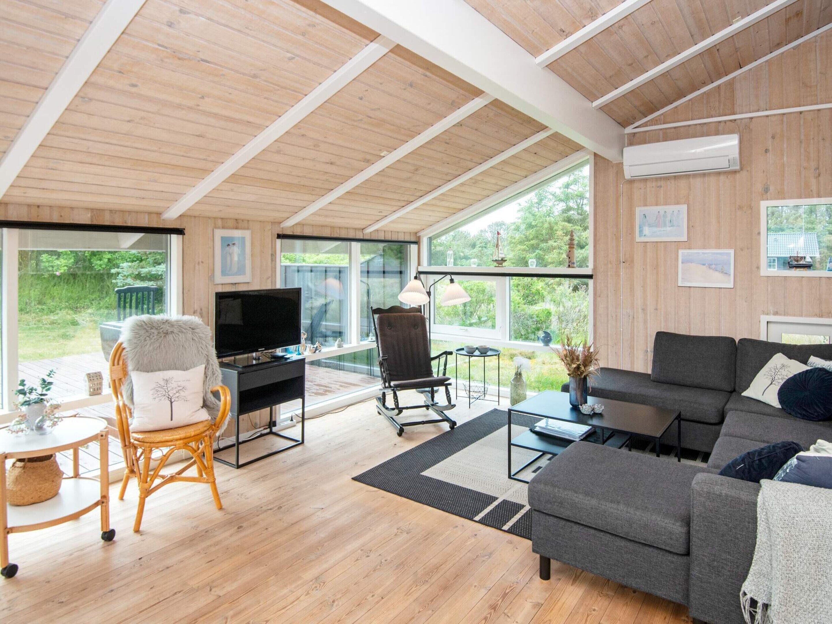 A modern living room with a wooden ceiling, large windows, a grey sofa, a TV, and a wicker chair.