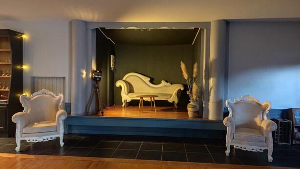 A room with a stage, two ornate chairs, a sofa, and a potted plant.