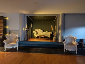 A room with a stage, two ornate chairs, a sofa, and a potted plant. - Hotel de la Plage (Le Nouvion-en-Thiérache)