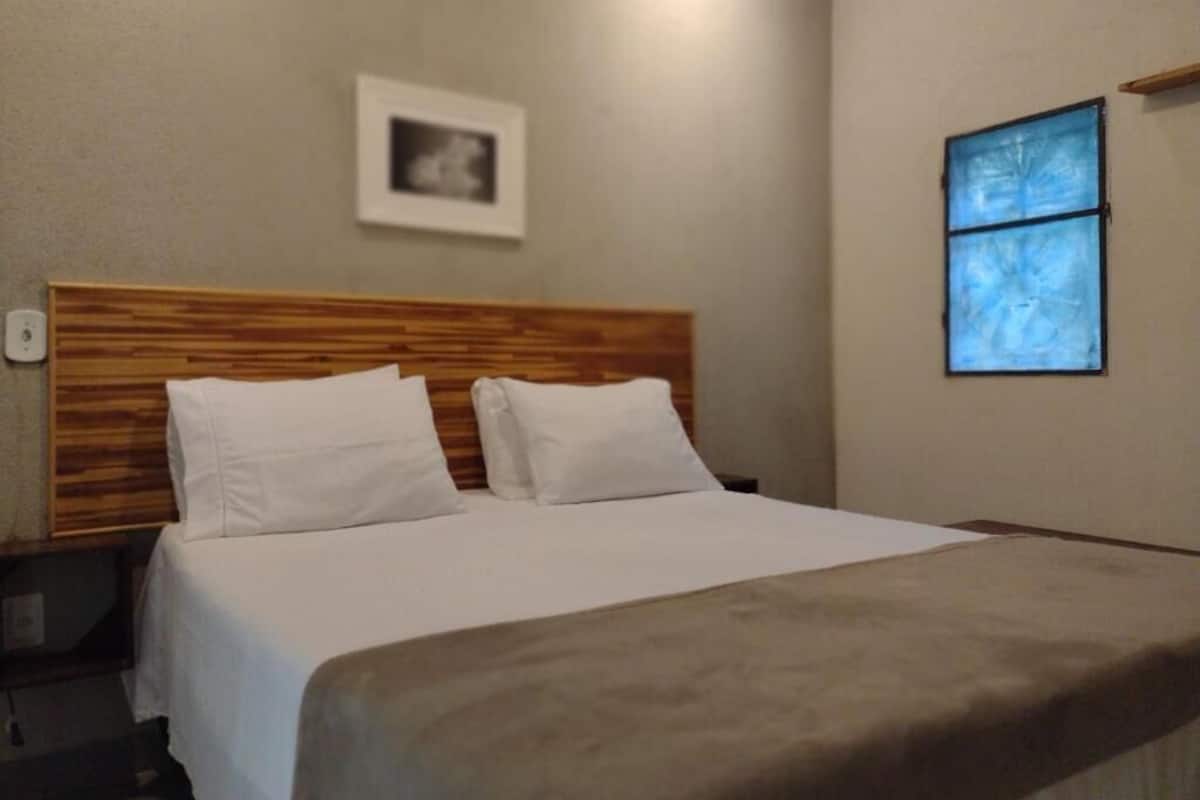 Una habitación de hotel con cabecera de madera, cama con ropa de cama blanca, ventana con vistas y un cuadro en la pared.